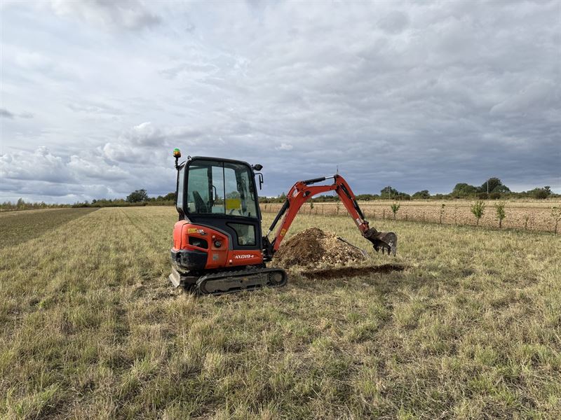Mini excavator opening a trial pit for BRE 365 infiltration testing in an open field